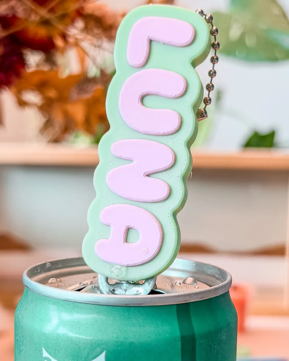 A mint-green can opener reading LUNA in pink letters, lifting the tab on a drinks can